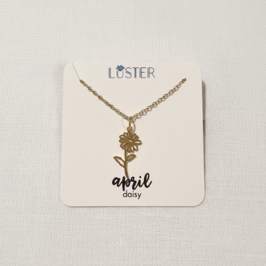 A gold-tone floral necklace on a delicate chain, displayed on a cream-colored Luster brand card that reads 'April Daisy', set against a light gray textured background.