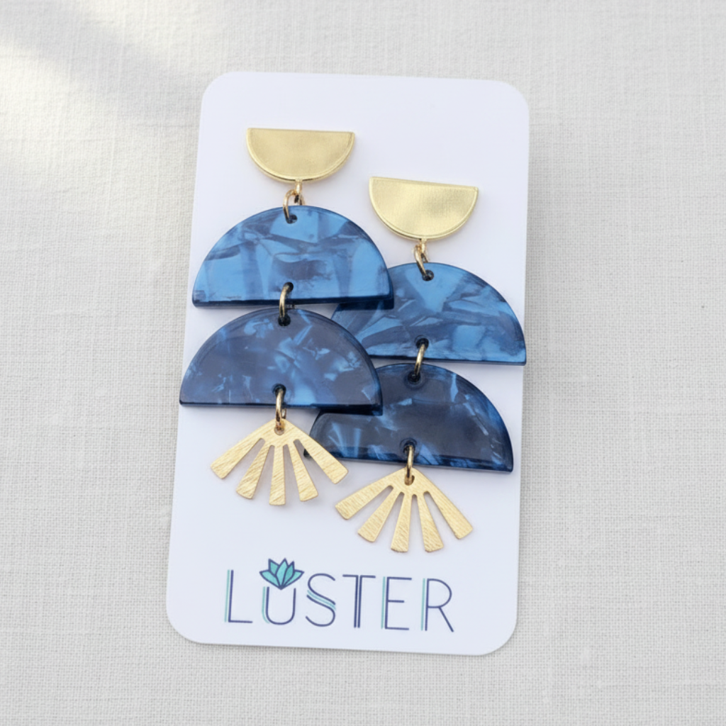 A pair of blue marbled geometric drop earrings with gold-tone accents, displayed on a white Luster brand card against a light textured fabric background.