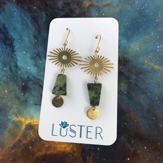 Pair of celestial-shaped earrings with green stones on a Luster branded card against a cosmic background
