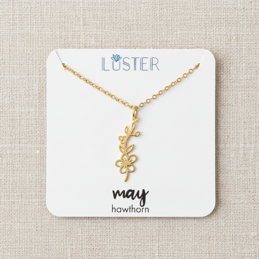 A gold hawthorn flower necklace on a delicate chain, presented on a white display card labeled 'LUSTER' and 'may hawthorn', set against a textured beige linen background.