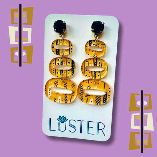 Pair of yellow and black patterned earrings on a Luster branded card against a purple background