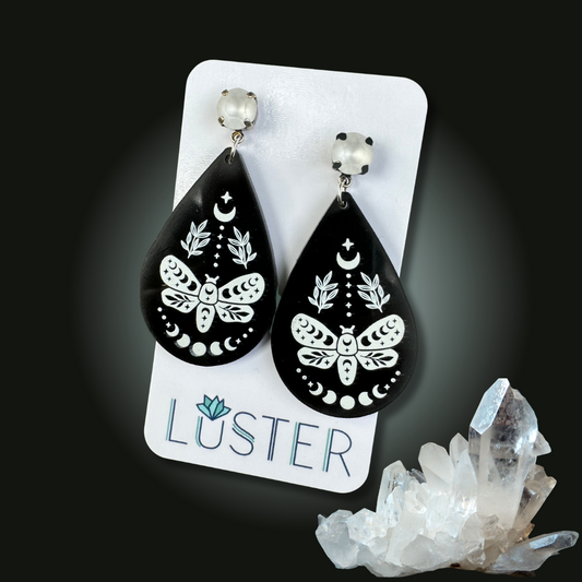 Black teardrop earrings with white butterfly design on a 'Luster' branded card, with crystals in the background.