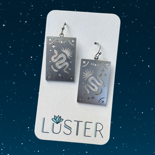 Pair of steel snake earrings on a Luster branded card against a starry night sky background