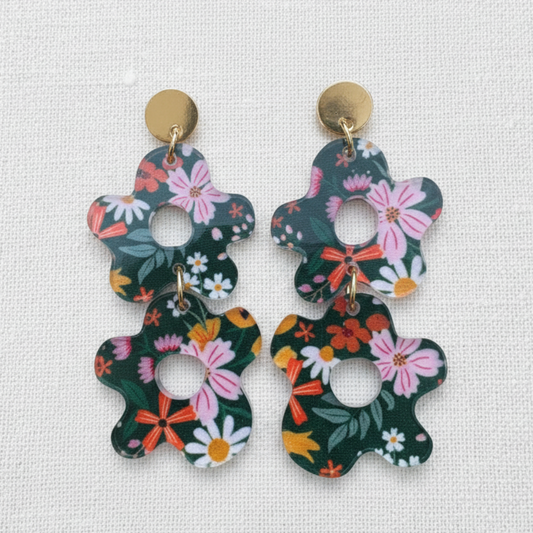 A pair of dangle earrings with a dark green floral pattern featuring pink, white, and orange flowers, suspended from gold-tone studs on a textured white fabric background.