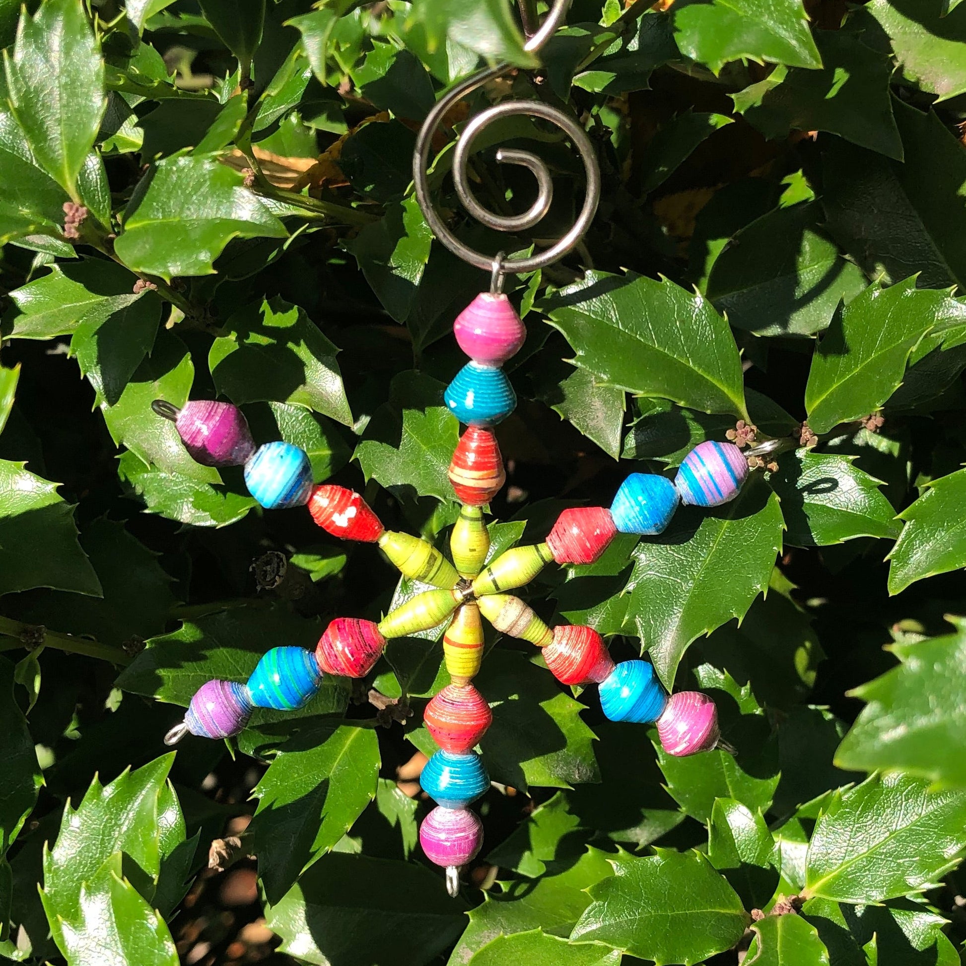 A colorful paper ornament with a star-shaped design, featuring multiple Fairtrade beads in blue, red, green, and yellow, and a metal hook at the top.