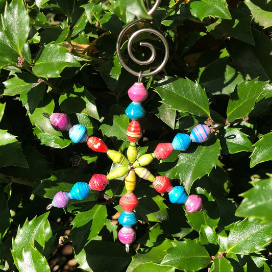 A colorful paper ornament with a star-shaped design, featuring multiple Fairtrade beads in blue, red, green, and yellow, and a metal hook at the top.