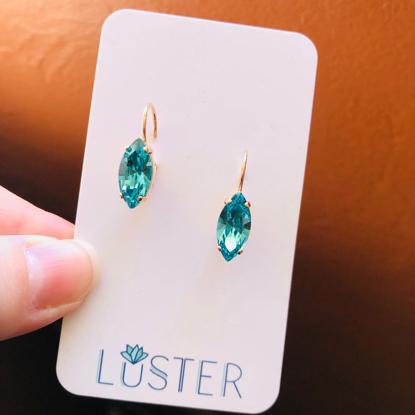 A pair of teal crystal navette leverback earrings displayed on a card with the brand name 'LUSTER'.